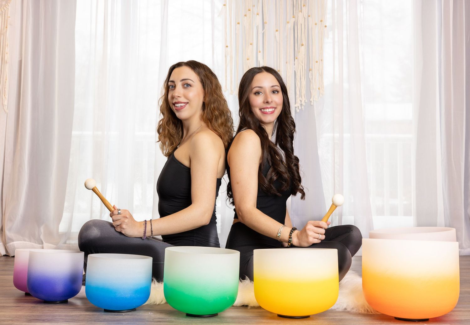 Two people sit back-to-back on a yoga mat with colorful crystal singing bowls arranged in front of them.