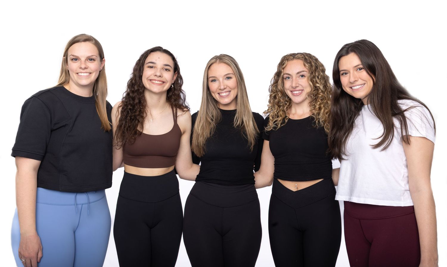 A group of young people stand together in a row wearing casual clothing in dark and neutral colors.
