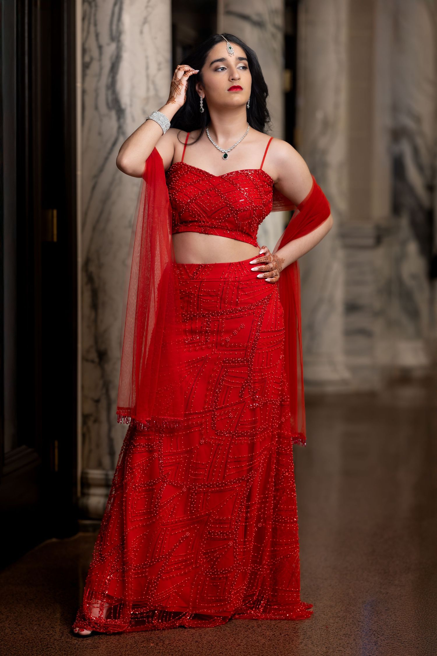 Woman in sparkly red evening gown with flowing tulle overlay posing against dark backdrop.