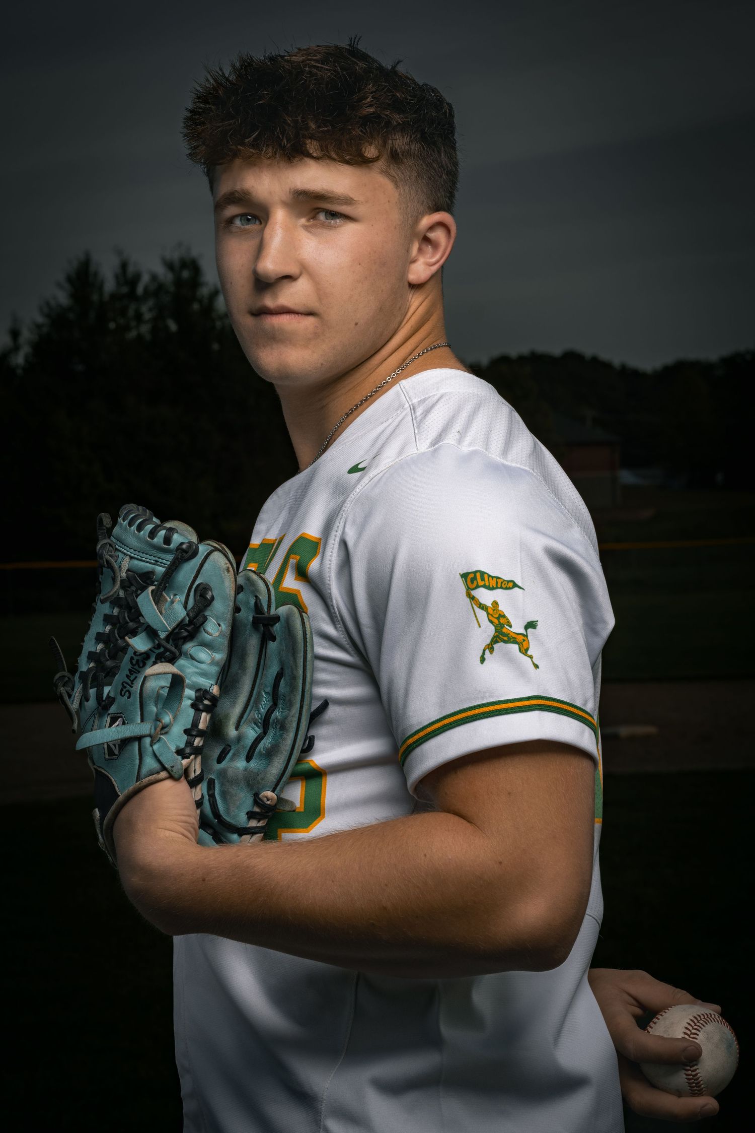 Baseball player in white uniform with team logo patch holds turquoise glove and ball in dramatic lighting.