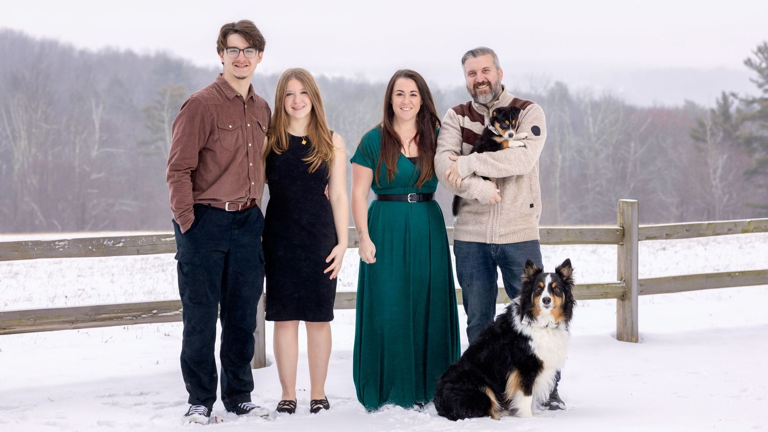 A family group poses together with their Bernese Mountain Dog on a snowy winter day by a wooden fence.