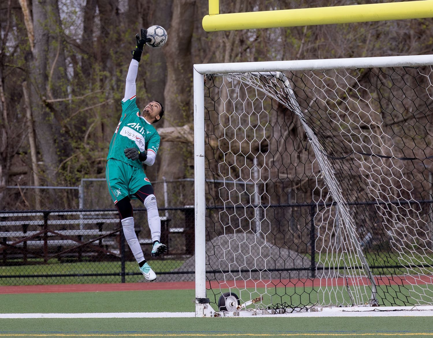 A soccer goalkeeper in a teal uniform makes a diving save to block a shot during a match.