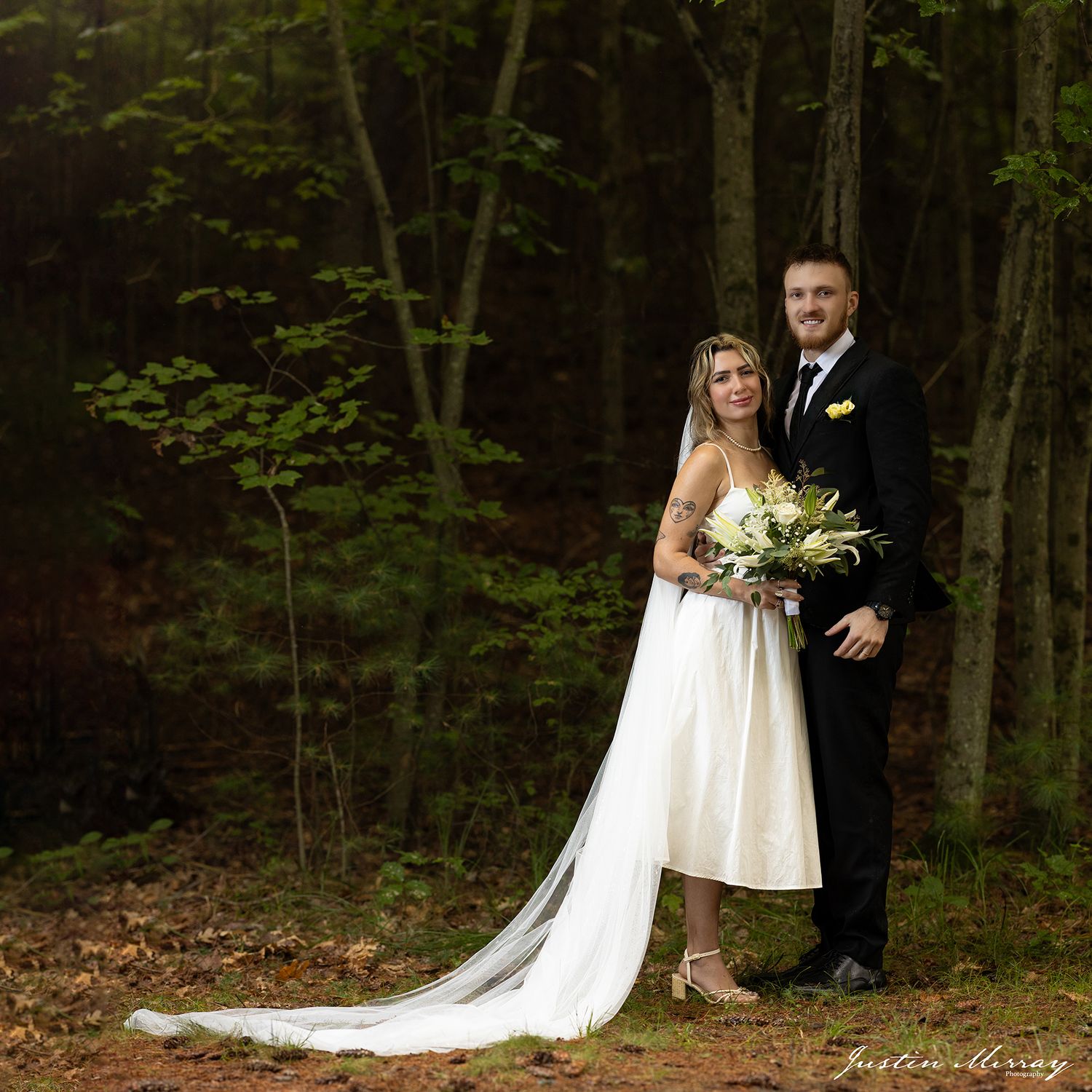 A couple in wedding attire poses together in a wooded forest setting with a long flowing white dress and bouquet.