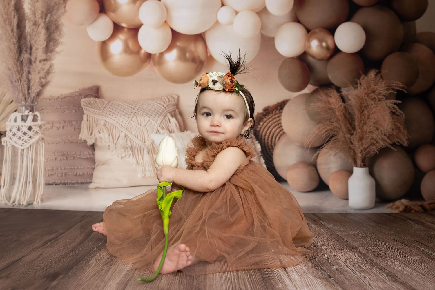 A baby in a brown tutu dress sits in front of a backdrop of neutral-colored balloons for a portrait session.