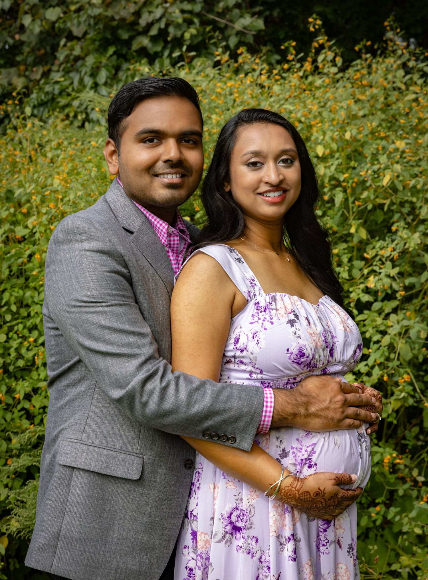 A couple poses together outdoors against a backdrop of yellow flowers and greenery wearing purple and grey attire.
