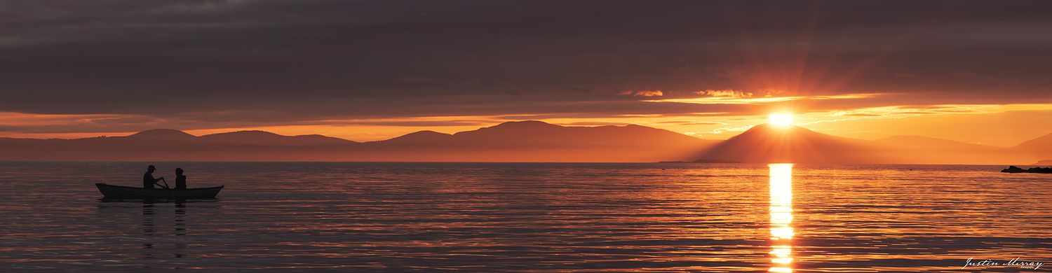 A dramatic sunset creates a golden reflection on calm ocean waters with silhouetted mountains in the distance.