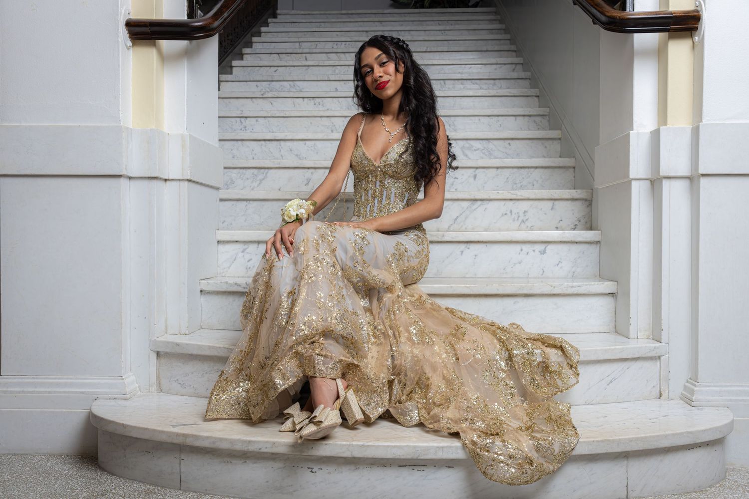 A person in a champagne-colored embroidered gown poses elegantly on white marble stairs in a glamorous photoshoot.