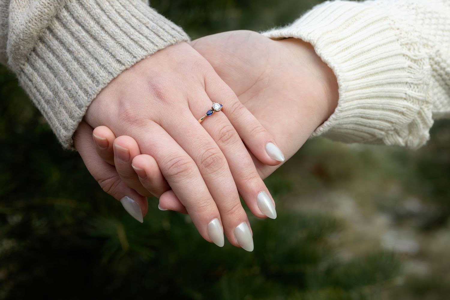 Two hands wearing cream sweaters touch tenderly while showing off an engagement ring.