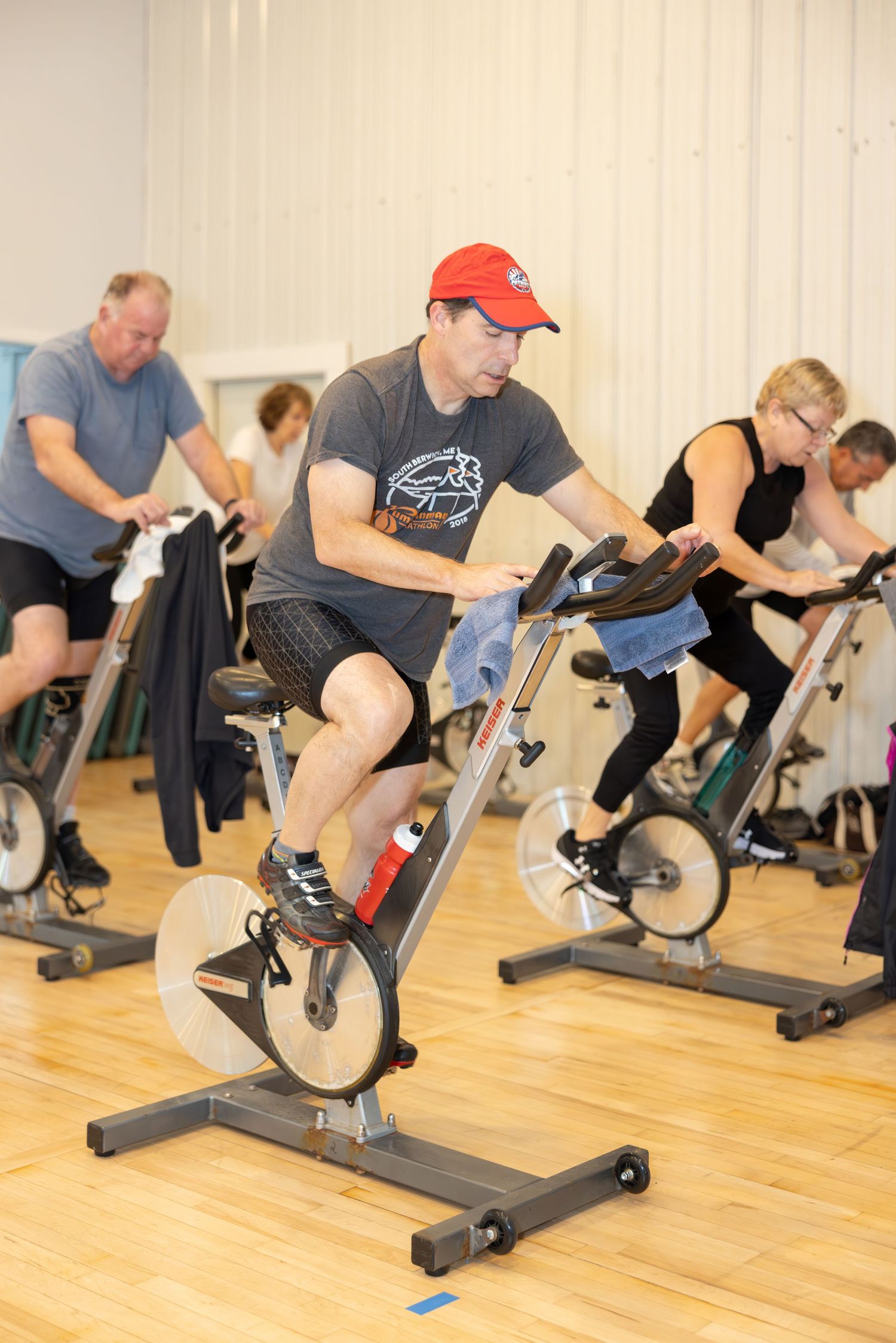 People exercising on stationary bikes during an indoor cycling class at a fitness center with wooden floors.