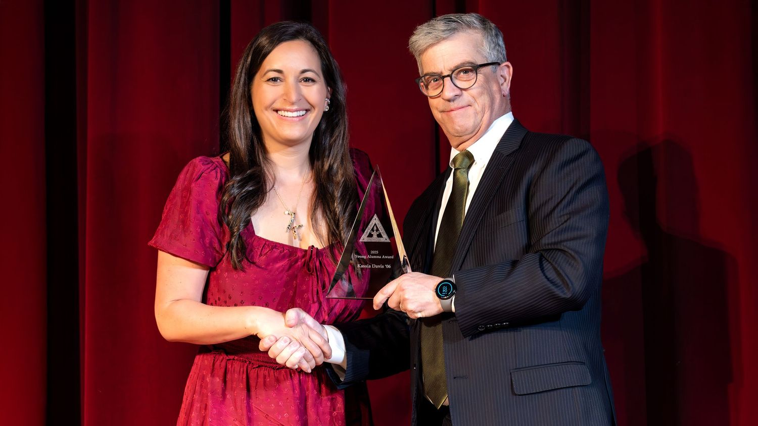 Two people in formal attire shake hands on stage while holding a crystal award trophy against a red curtain backdrop.