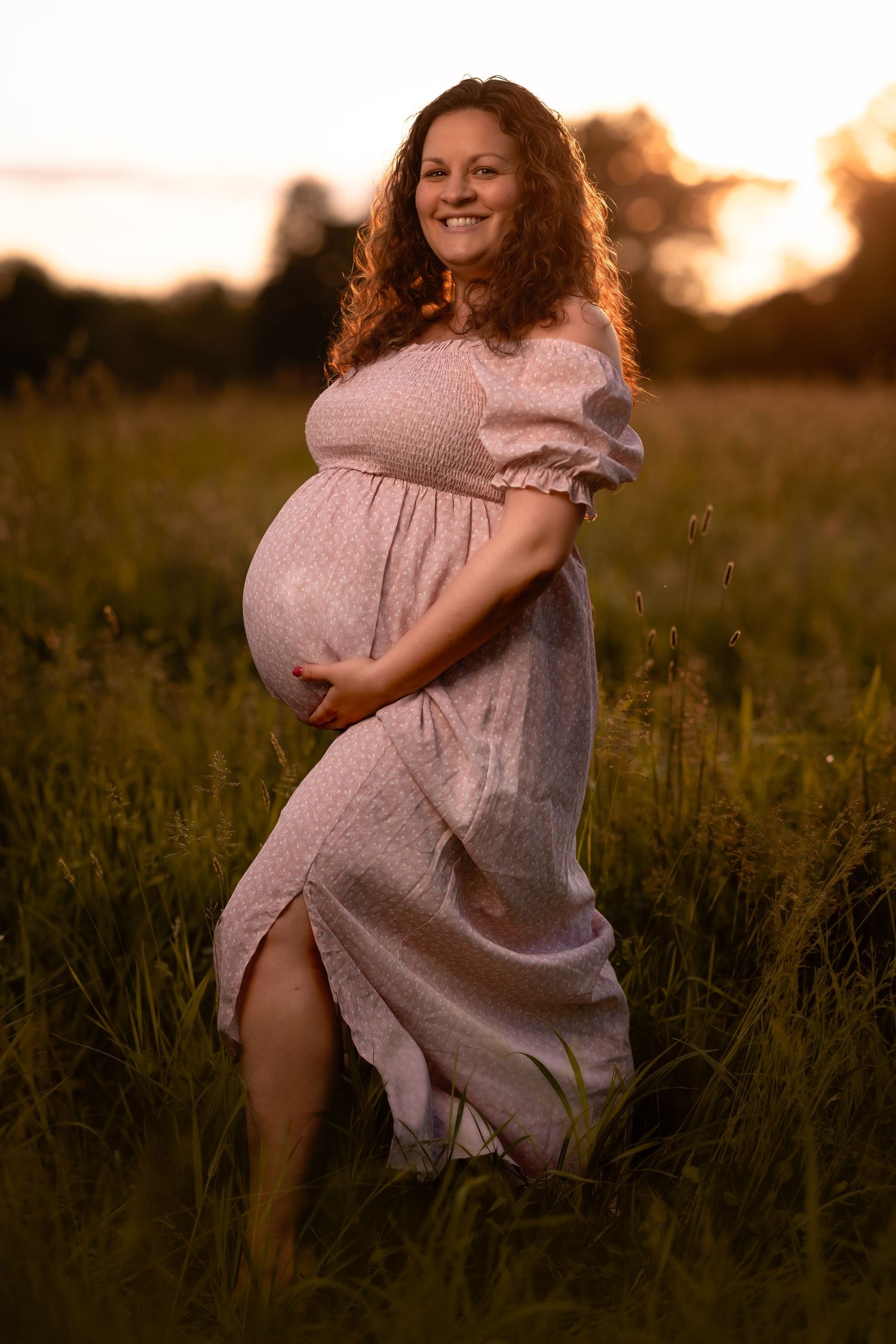 A pregnant person in a flowing pink dress poses in a golden field at sunset, creating a dreamy maternity portrait.