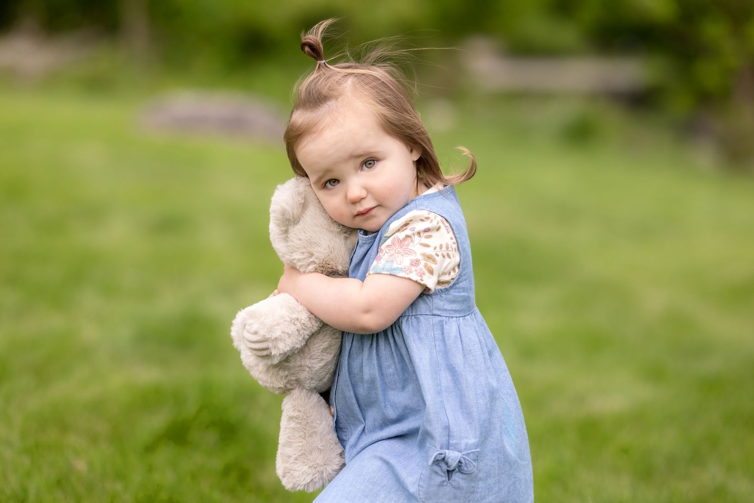 A young child in a denim dress hugs a large teddy bear while playing outdoors on green grass.