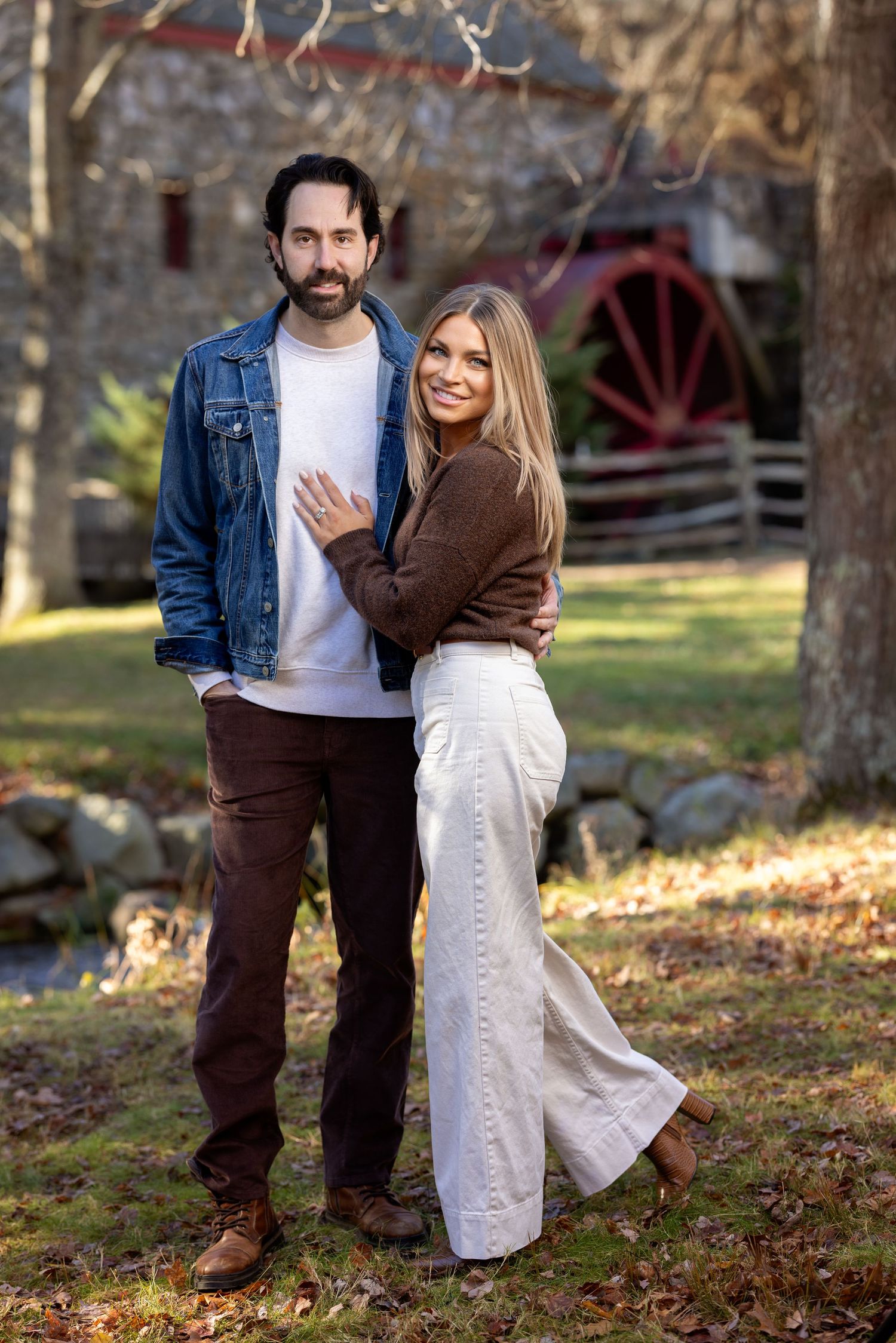 A couple embraces outdoors in autumn, wearing contrasting brown and white outfits against a rustic backdrop with fallen leaves.
