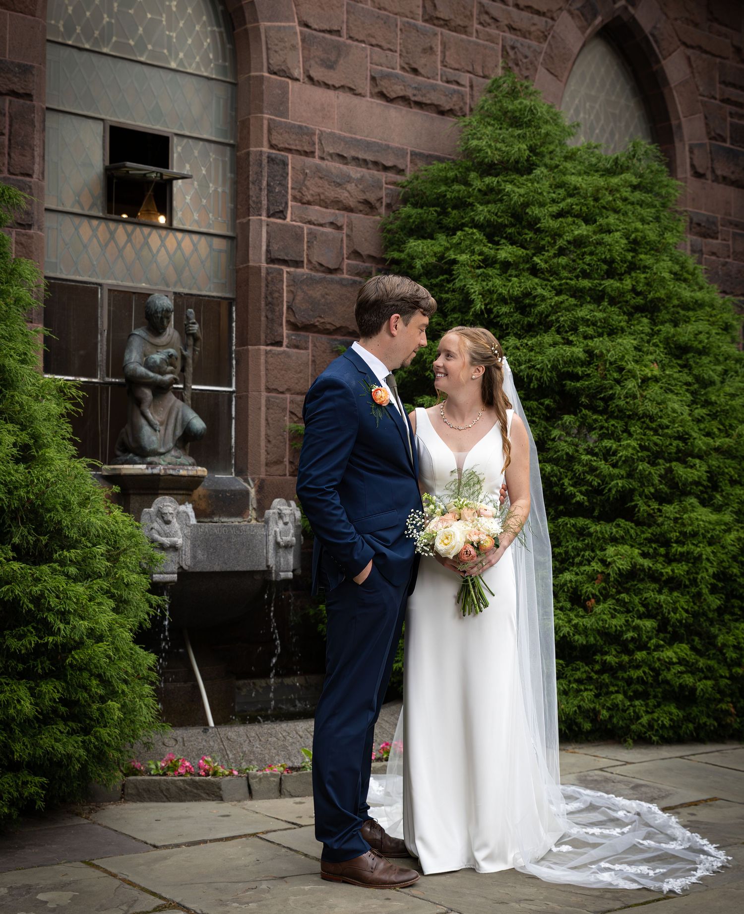 A couple in wedding attire share an intimate moment outside a stone church with arched windows and evergreen shrubs.