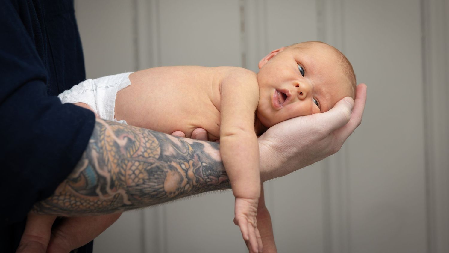 A newborn baby rests peacefully in tattooed arms against a white background.