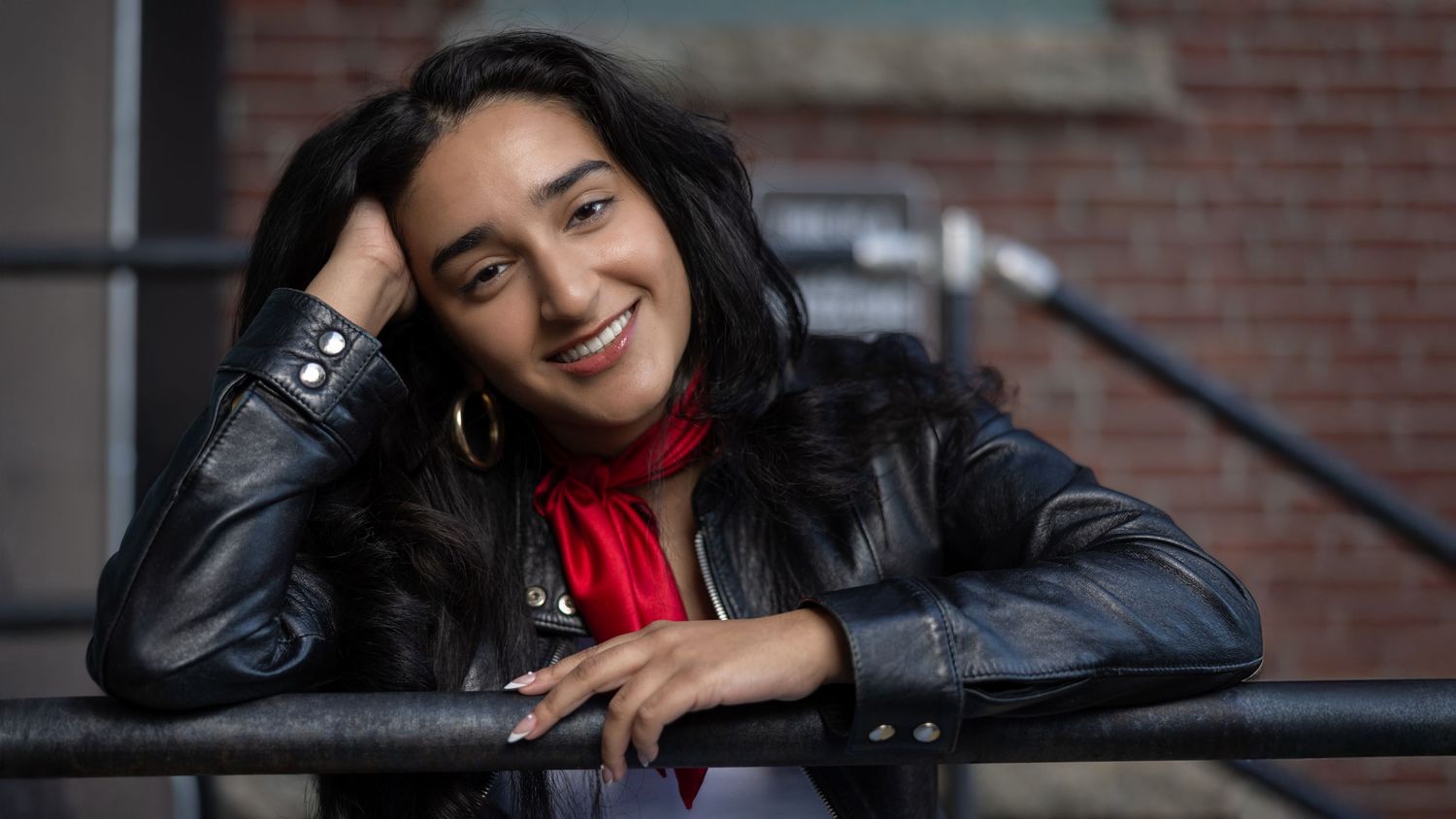 A person in a black leather jacket and red scarf poses playfully on a railing outdoors.