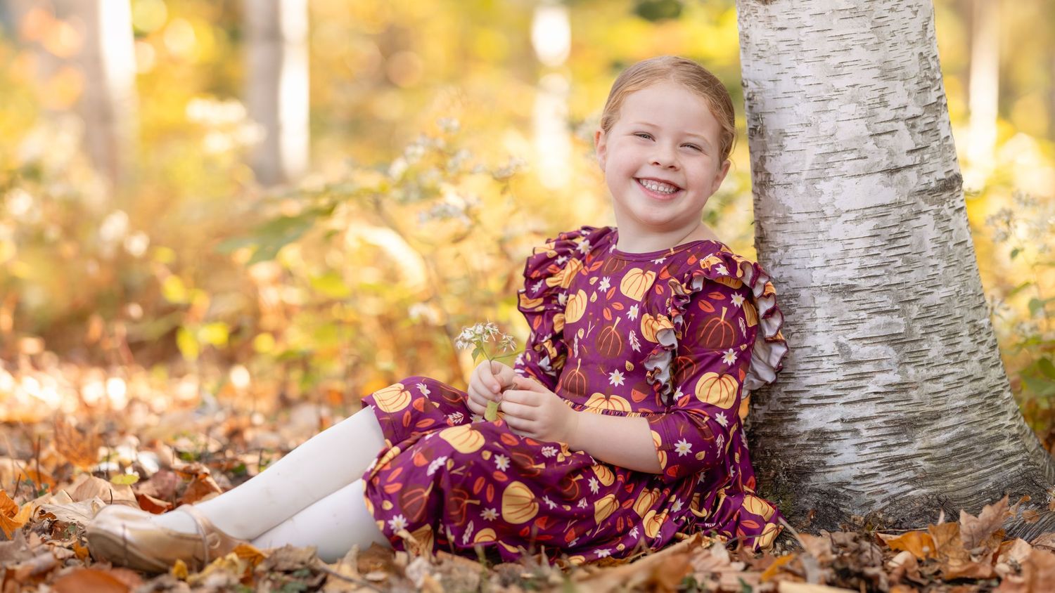 A person in a burgundy floral dress sits among autumn leaves near a birch tree trunk in golden sunlight.