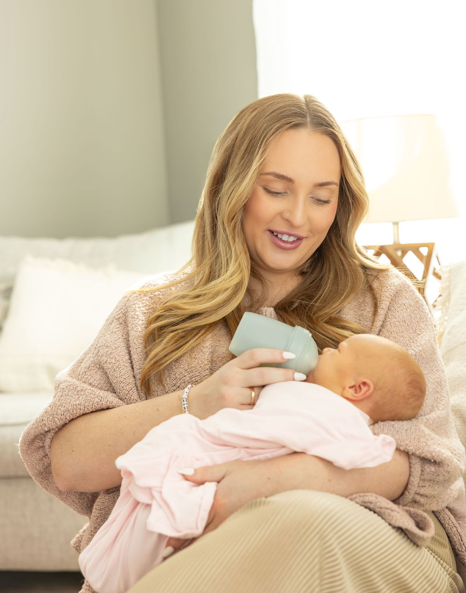 Parent in cozy sweater lovingly feeds and bonds with baby in pink outfit on soft beige couch.