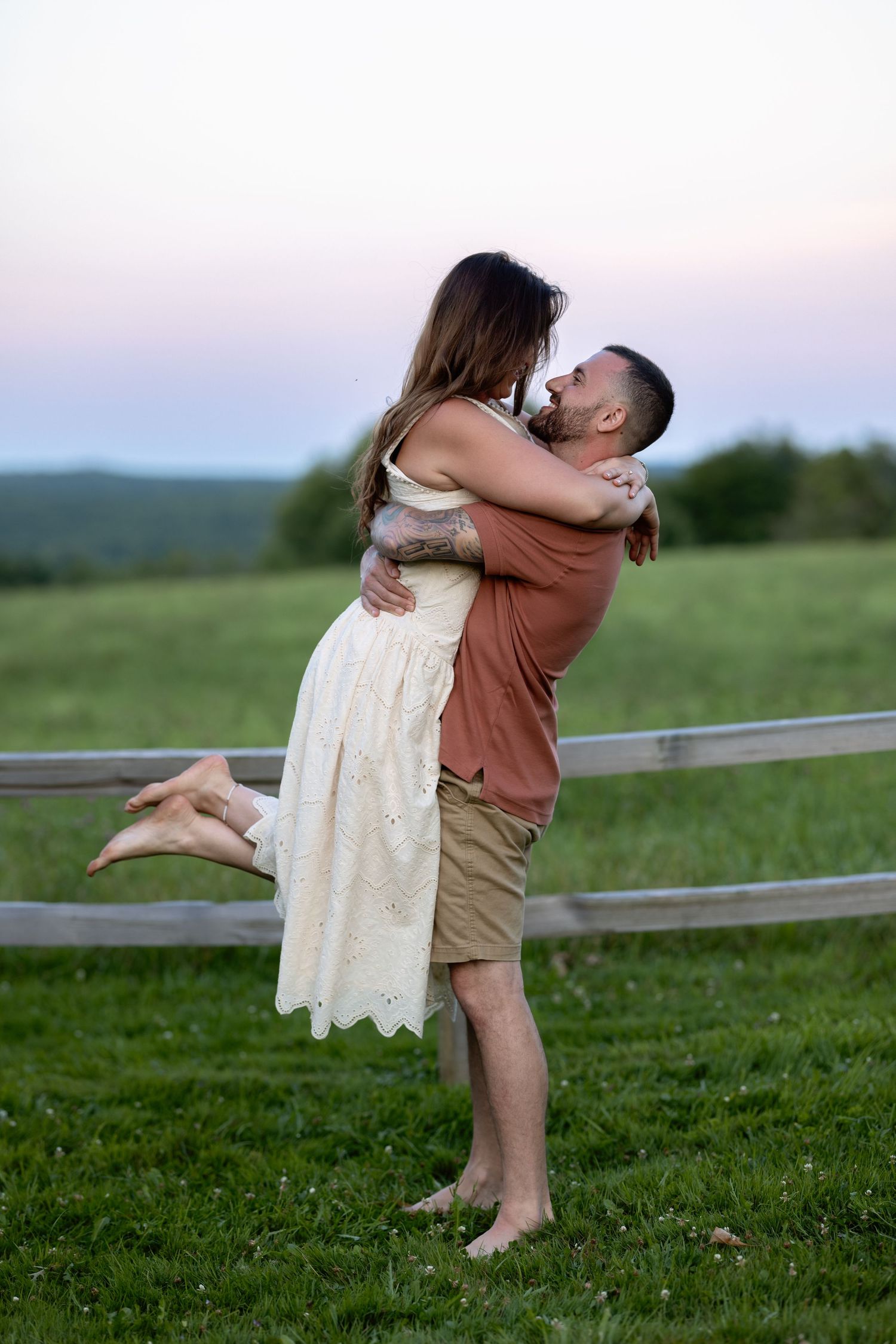 A romantic couple embraces in a grassy field at sunset with a wooden fence in the background.