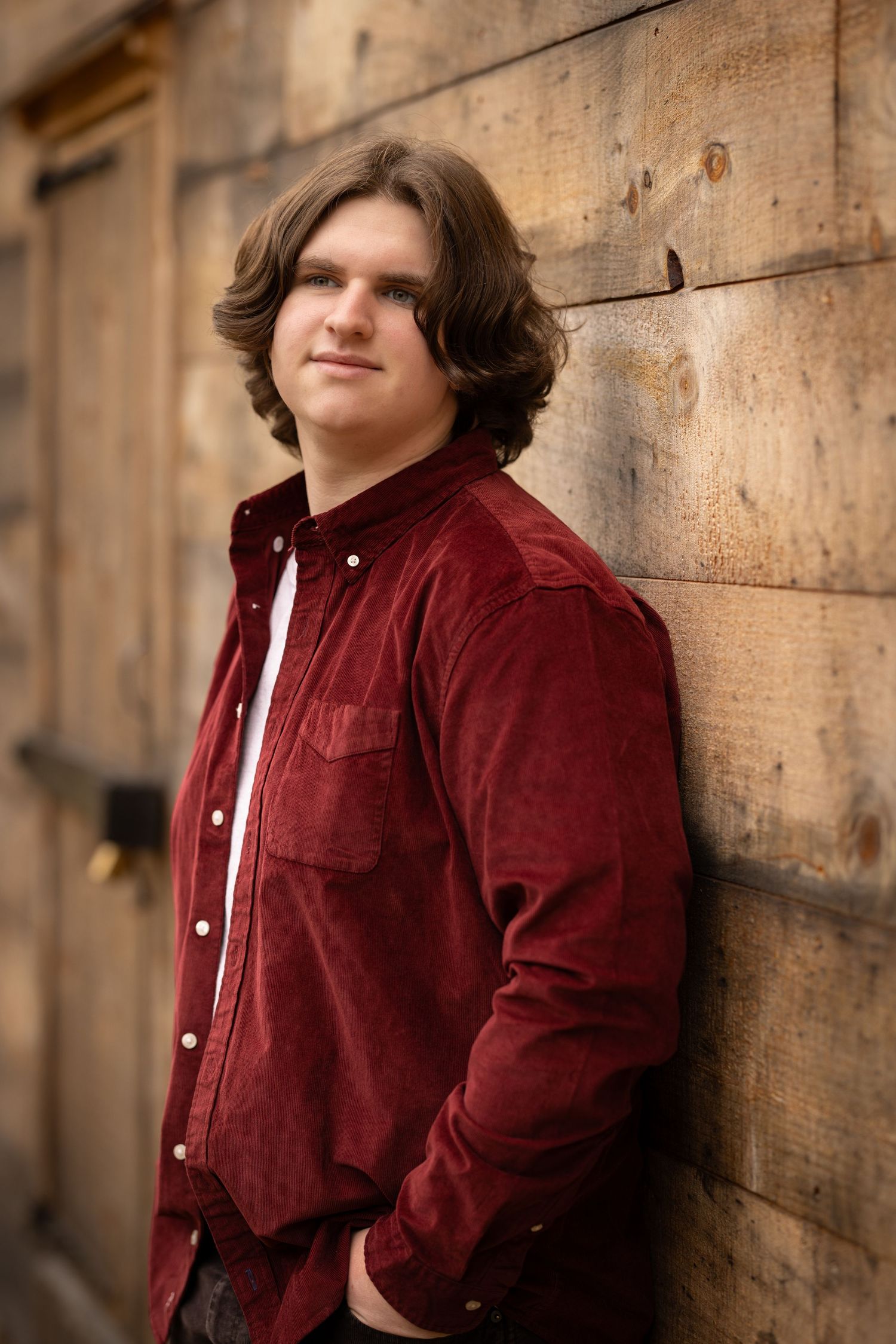 A person in a burgundy corduroy jacket leans against a wooden wall in dramatic lighting.