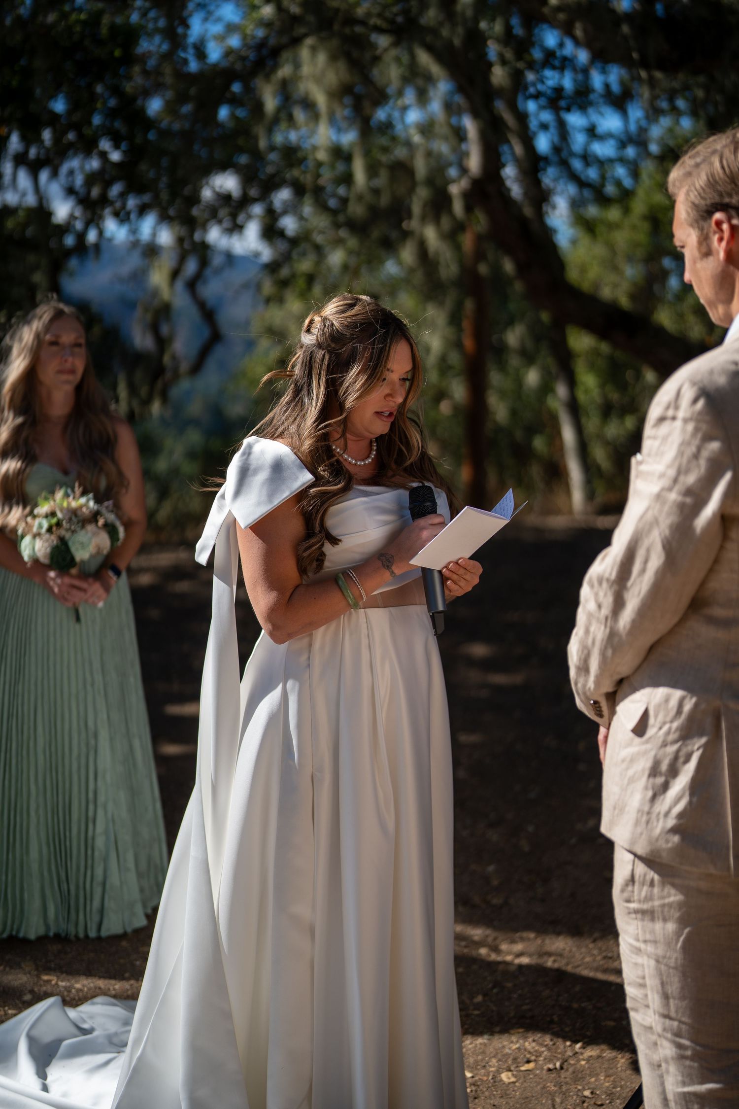 A bride in a flowing white dress reads vows during an outdoor wedding ceremony under trees.