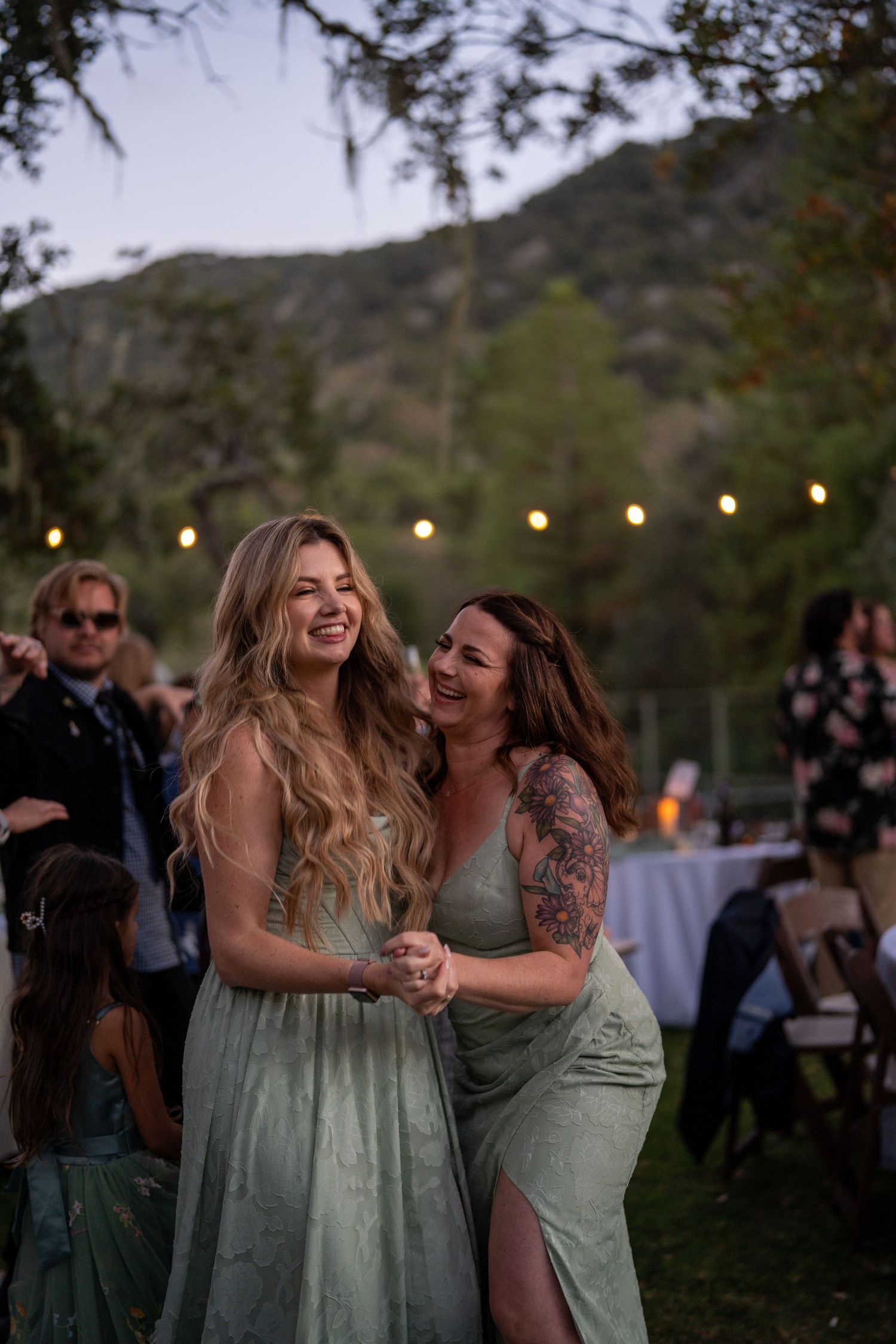 Friends share joyful laughter and embraces while dancing at an outdoor evening wedding reception with string lights.