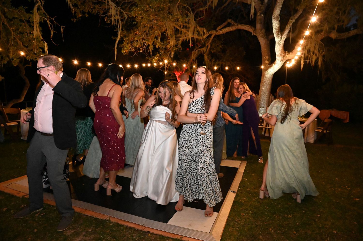 People dancing on a wooden dance floor at an outdoor evening party with trees illuminated in the background.