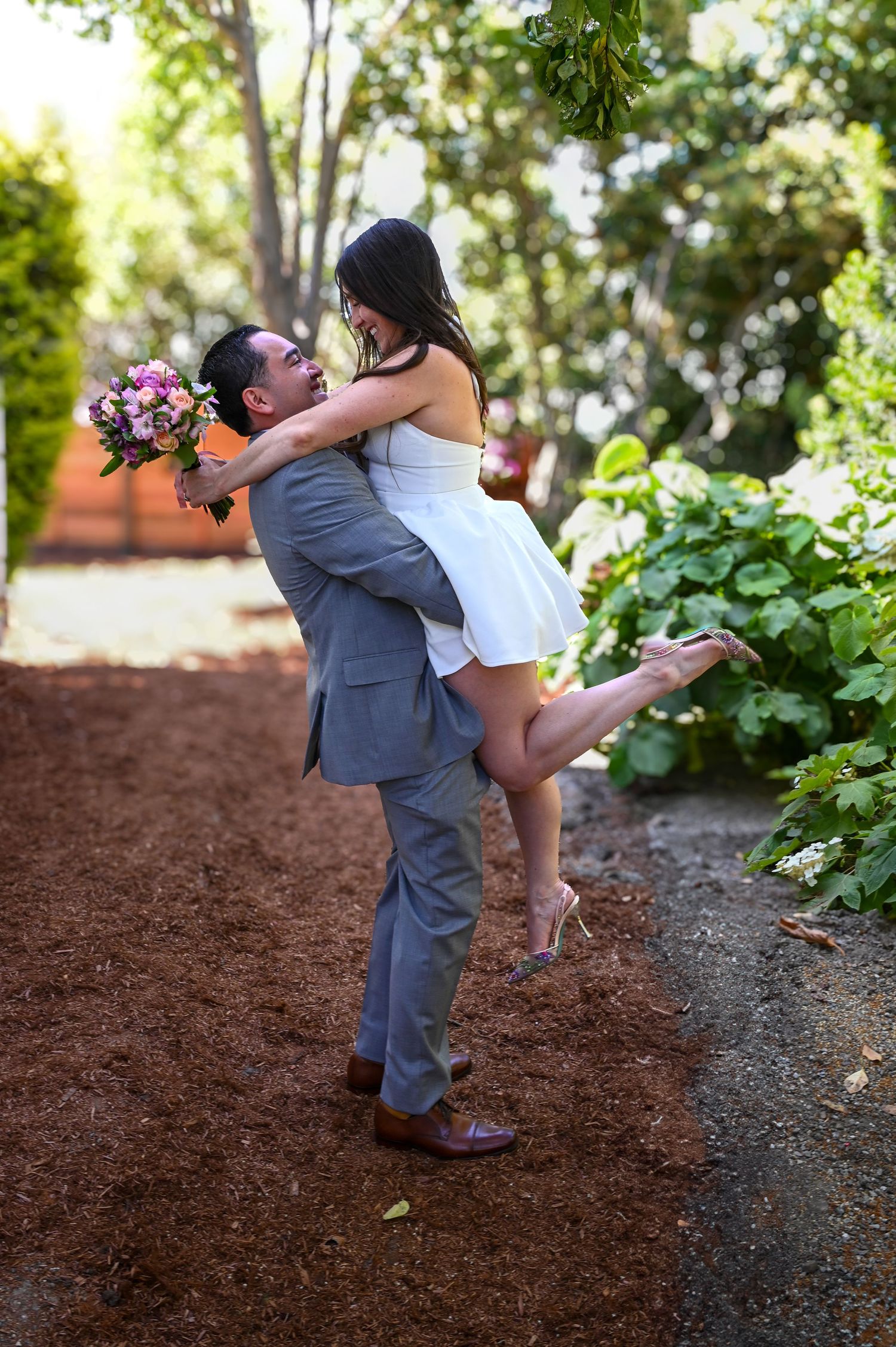 A playful dip kiss between a couple on a garden path surrounded by mulch and foliage.