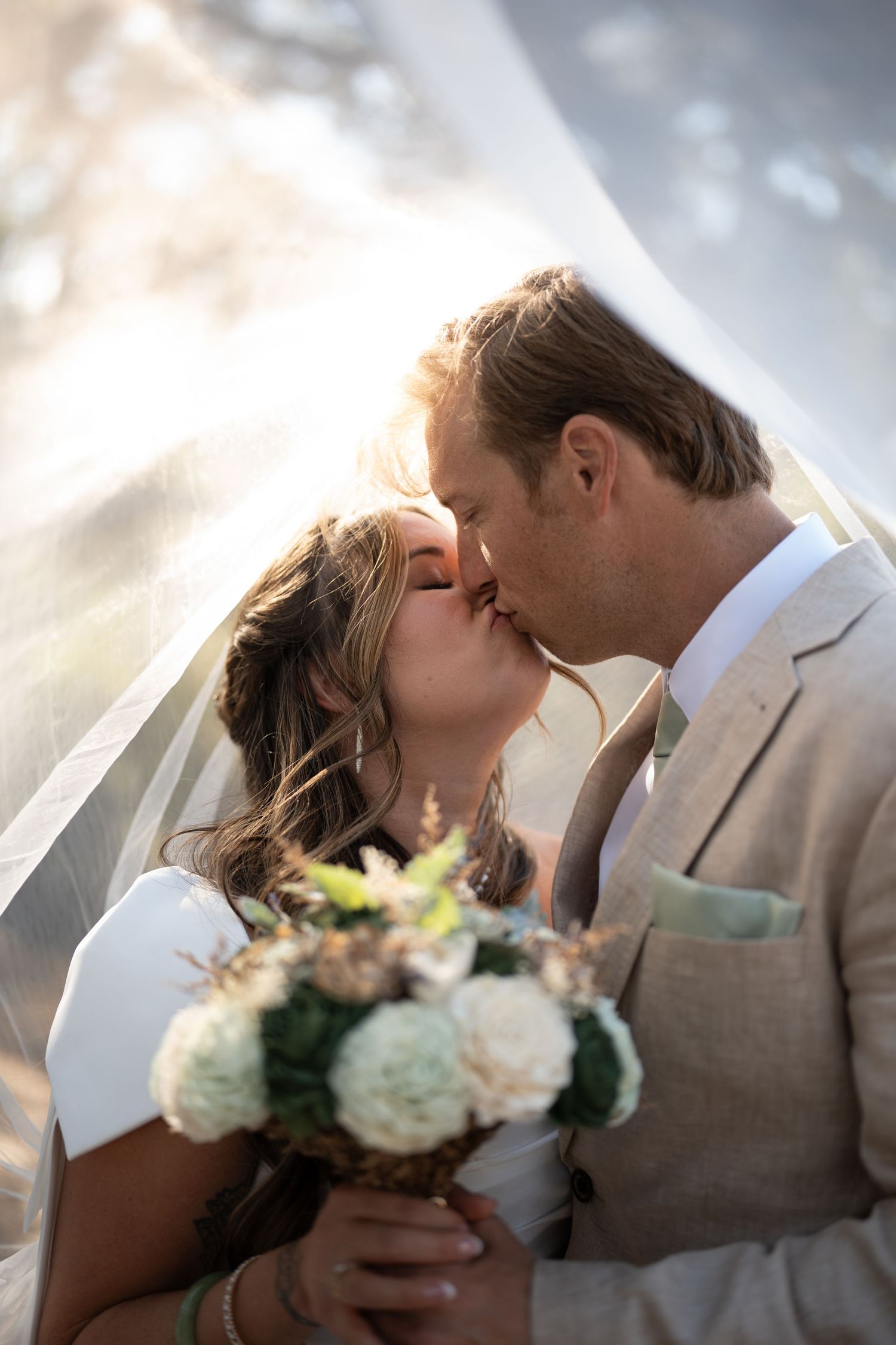 Romantic wedding kiss scene with couple in white dress and tan suit holding floral bouquet.