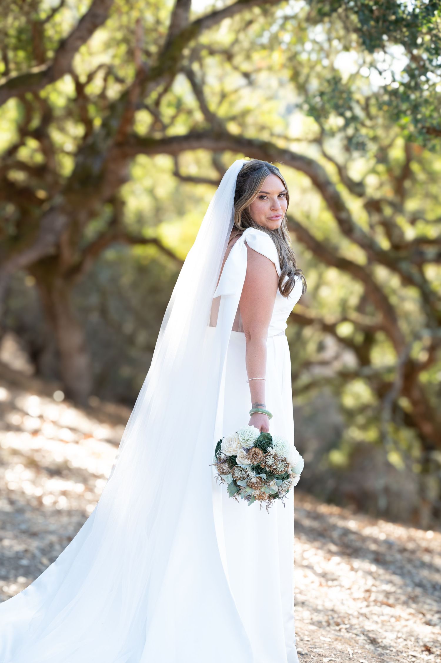 A bride in a white cap sleeve wedding gown and long veil poses with her bouquet in a sunlit wooded setting.