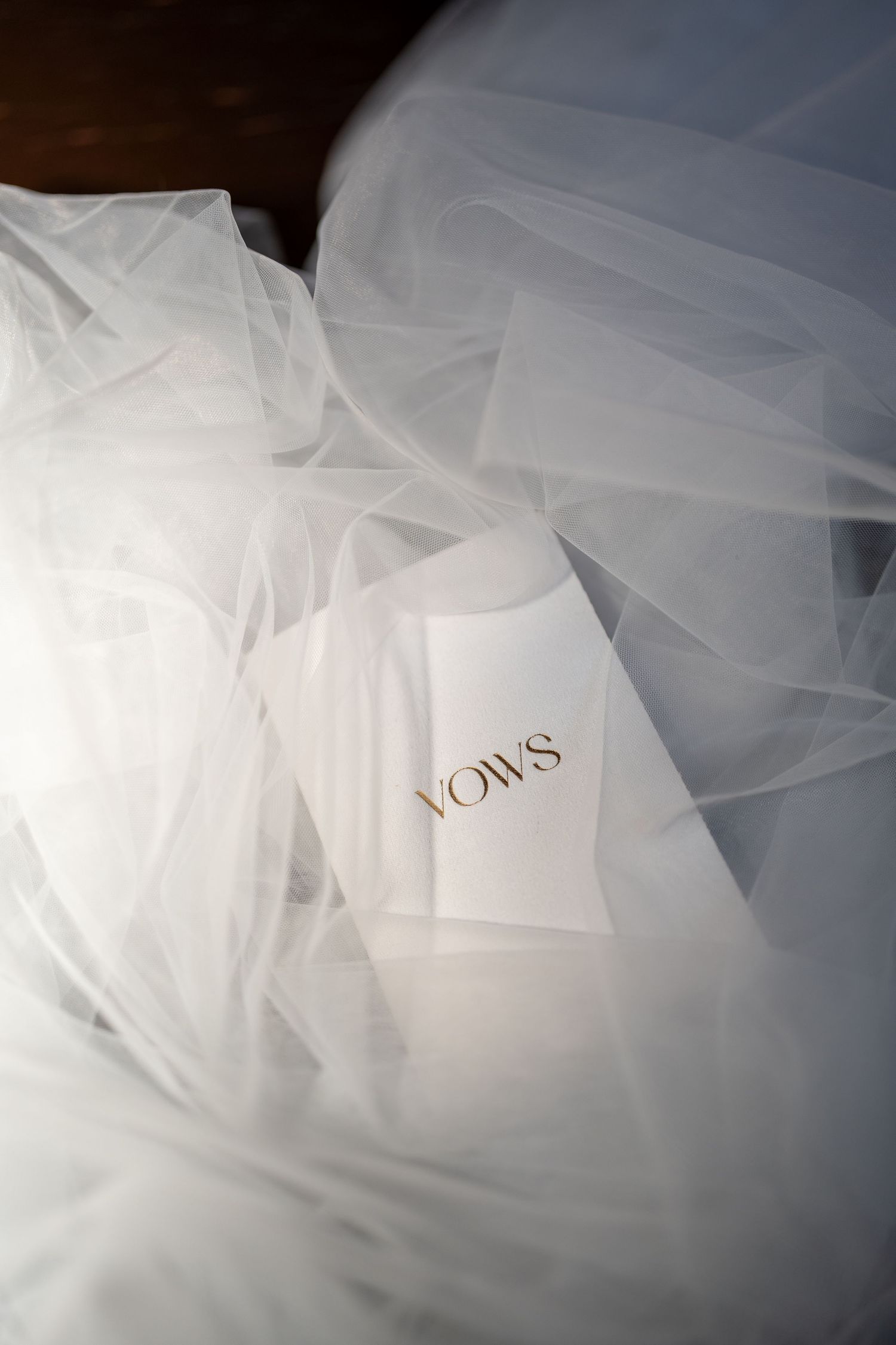 A delicate white wedding veil with the word 'vows' written in gold lettering lays softly against a light background.