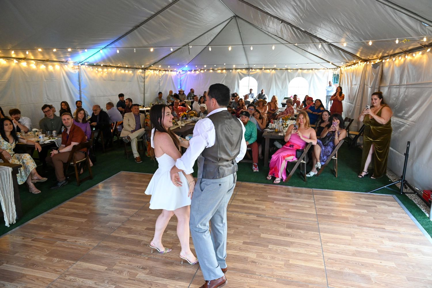 Wedding guests dance and celebrate on a wooden dance floor under a large white tent with string lights and decorations.