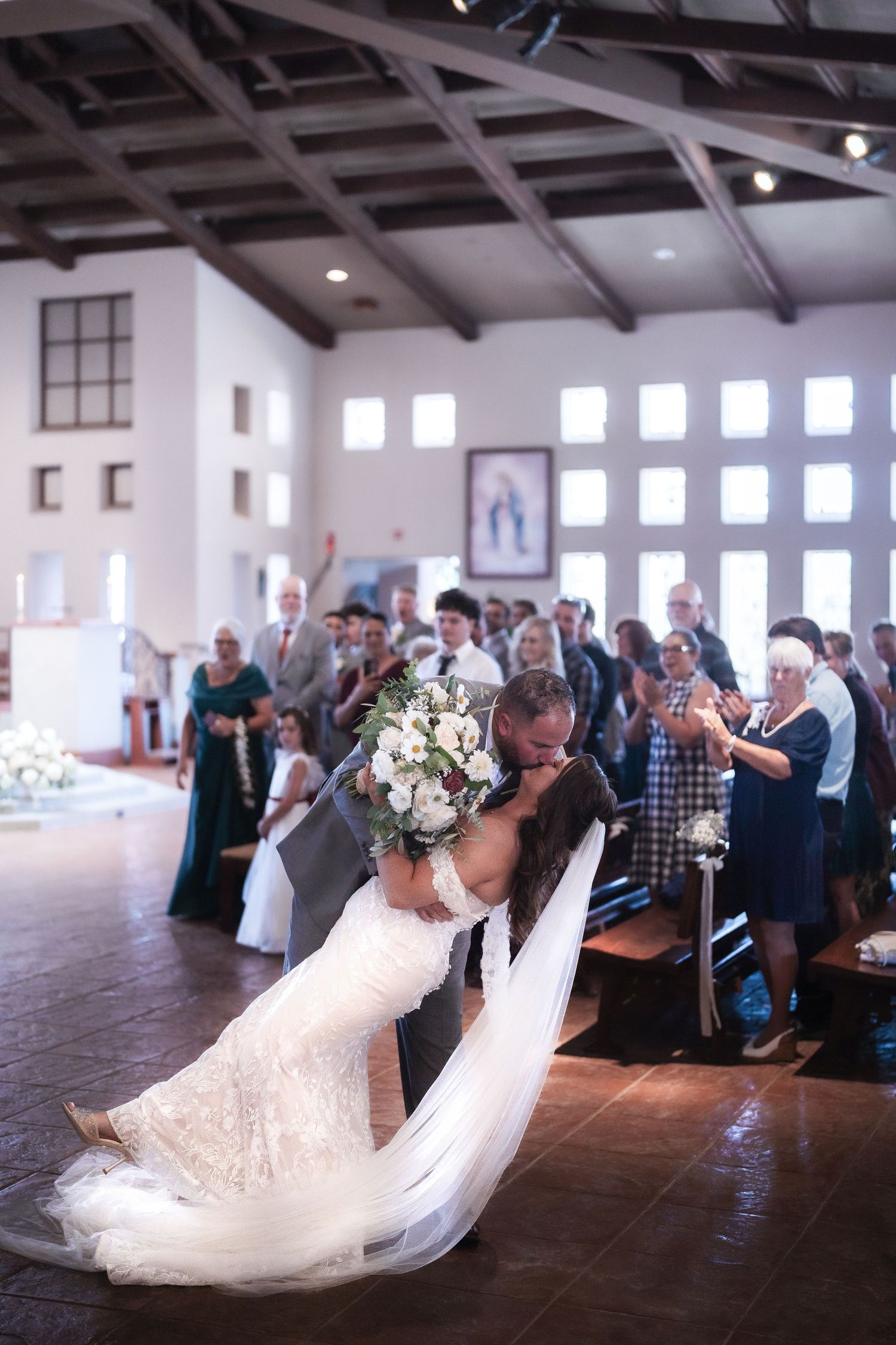 Wedding guests stand in rows during an emotional ceremony in a bright white room with wooden beams.