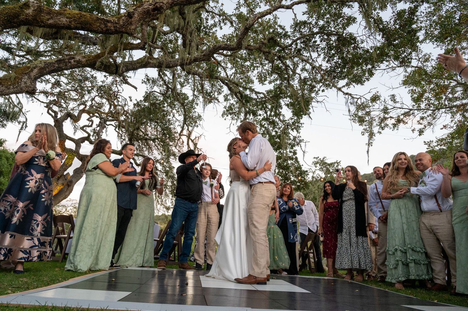 Wedding guests gather outdoors under a tree canopy during a sunset ceremony in a garden setting.