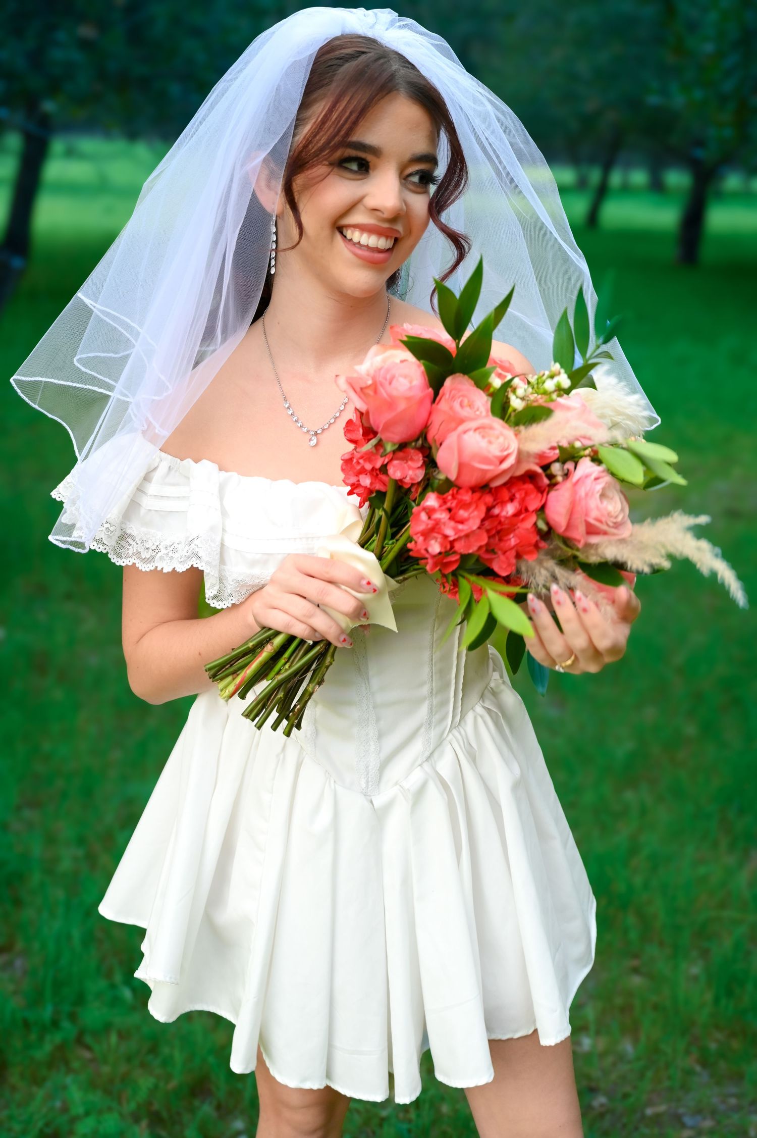 Bride wearing short white dress and veil smiles while holding pink and coral floral bouquet outdoors.