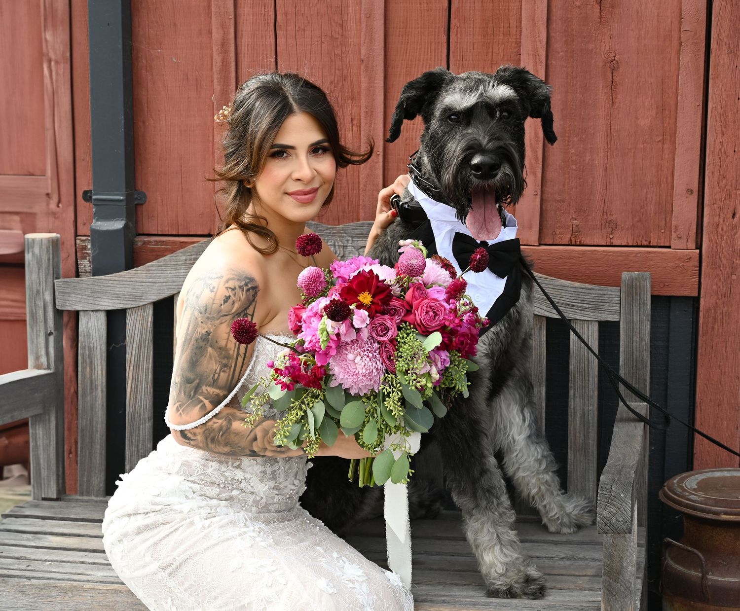 Bride in lace gown sits on bench with large black dog wearing bow tie tuxedo.
