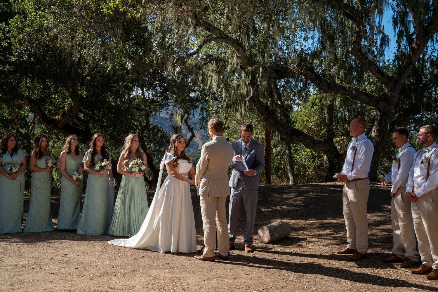 A wedding party dressed in white and beige poses together under shady trees in an outdoor garden setting.