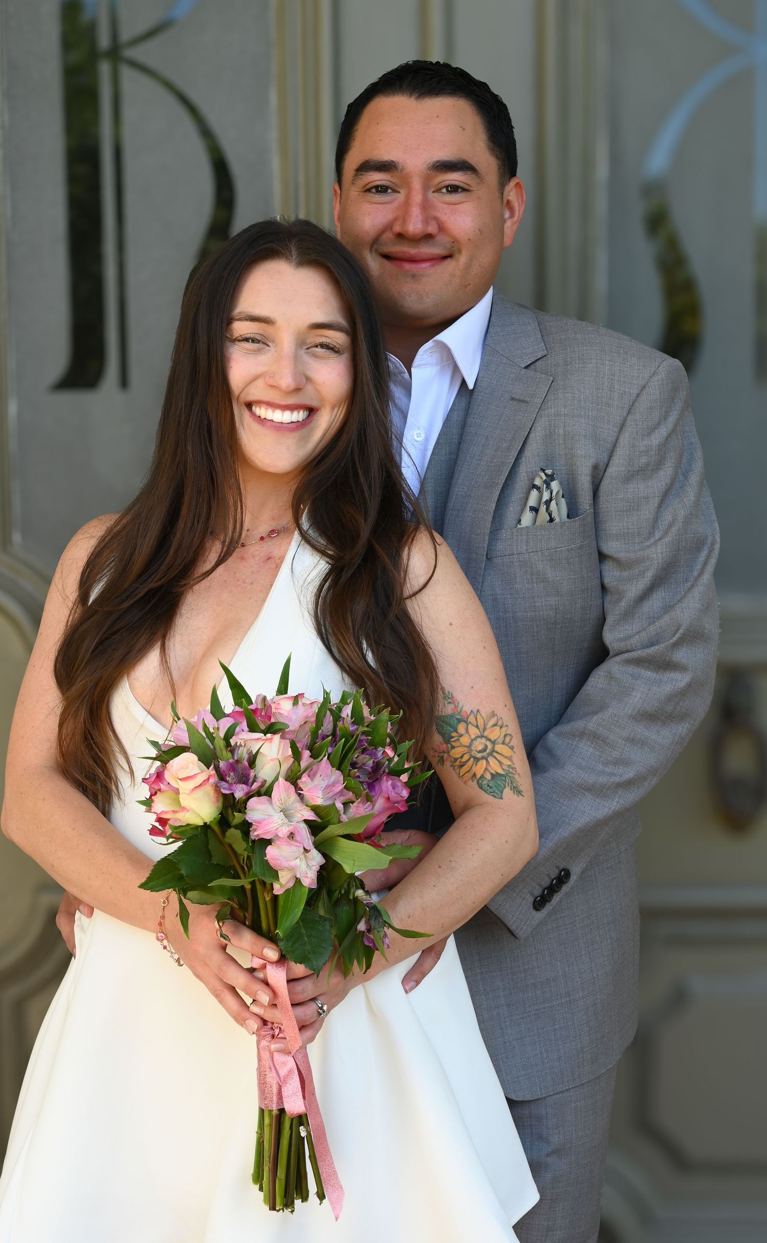Wedding couple poses together on porch with pink and purple bouquet.
