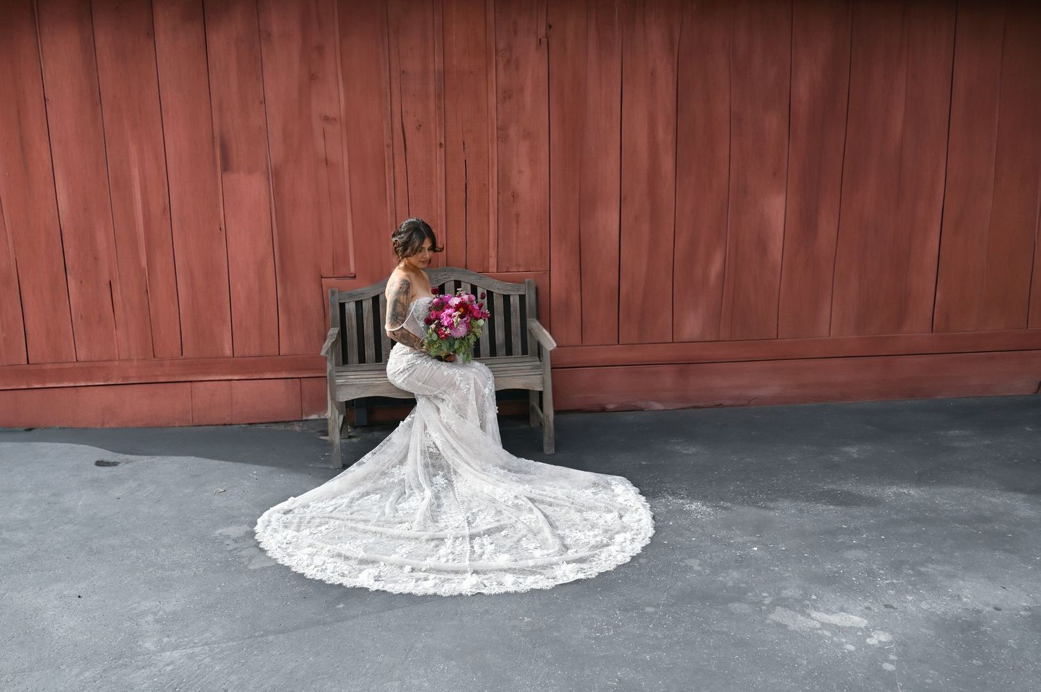 Bride in a white wedding dress with lace details sitting on a wooden floor against a rustic red barn wall.