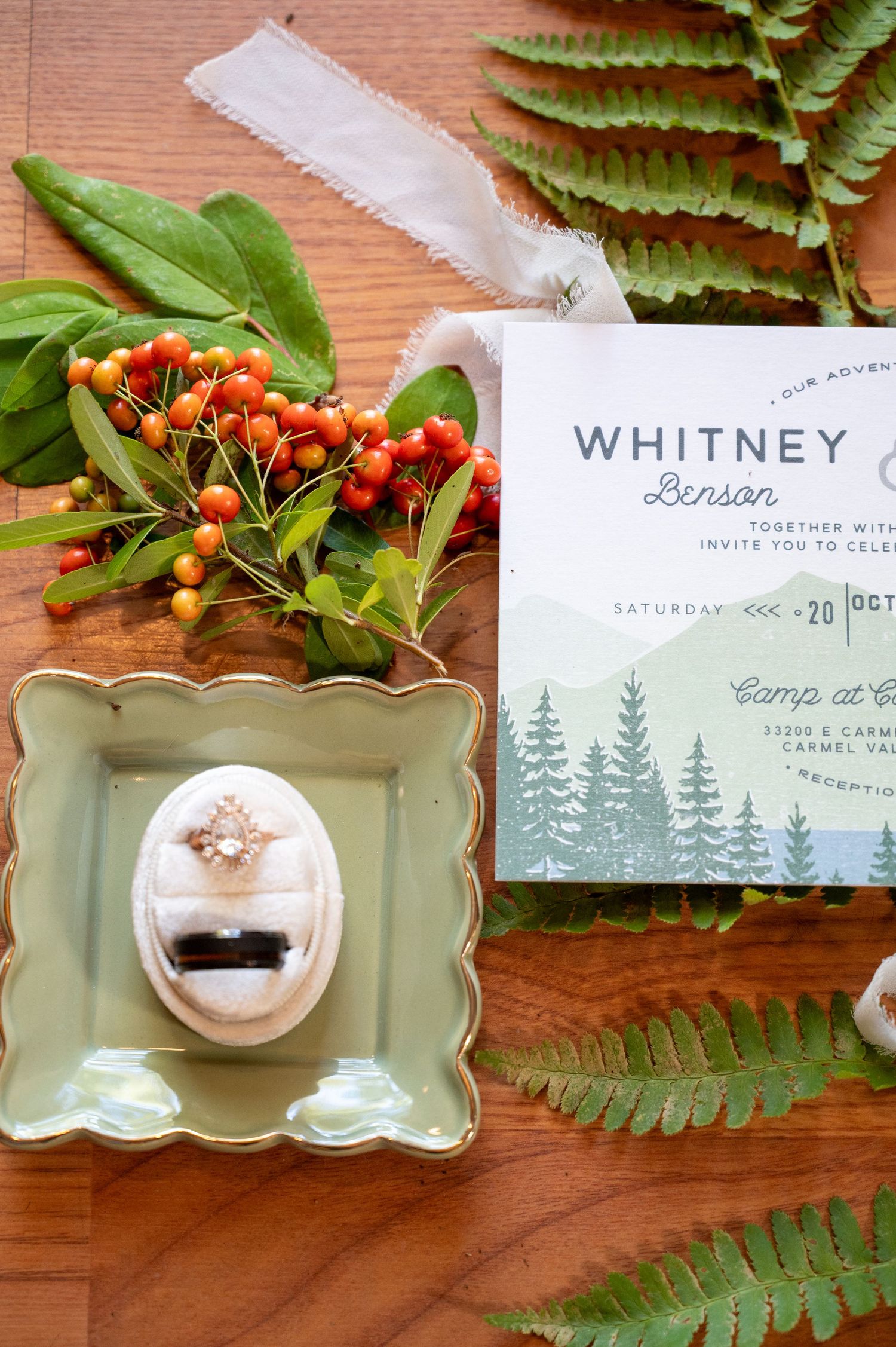 Wedding invitation with forest design displayed with ferns, orange berries, and a mint green decorative dish on wooden surface.