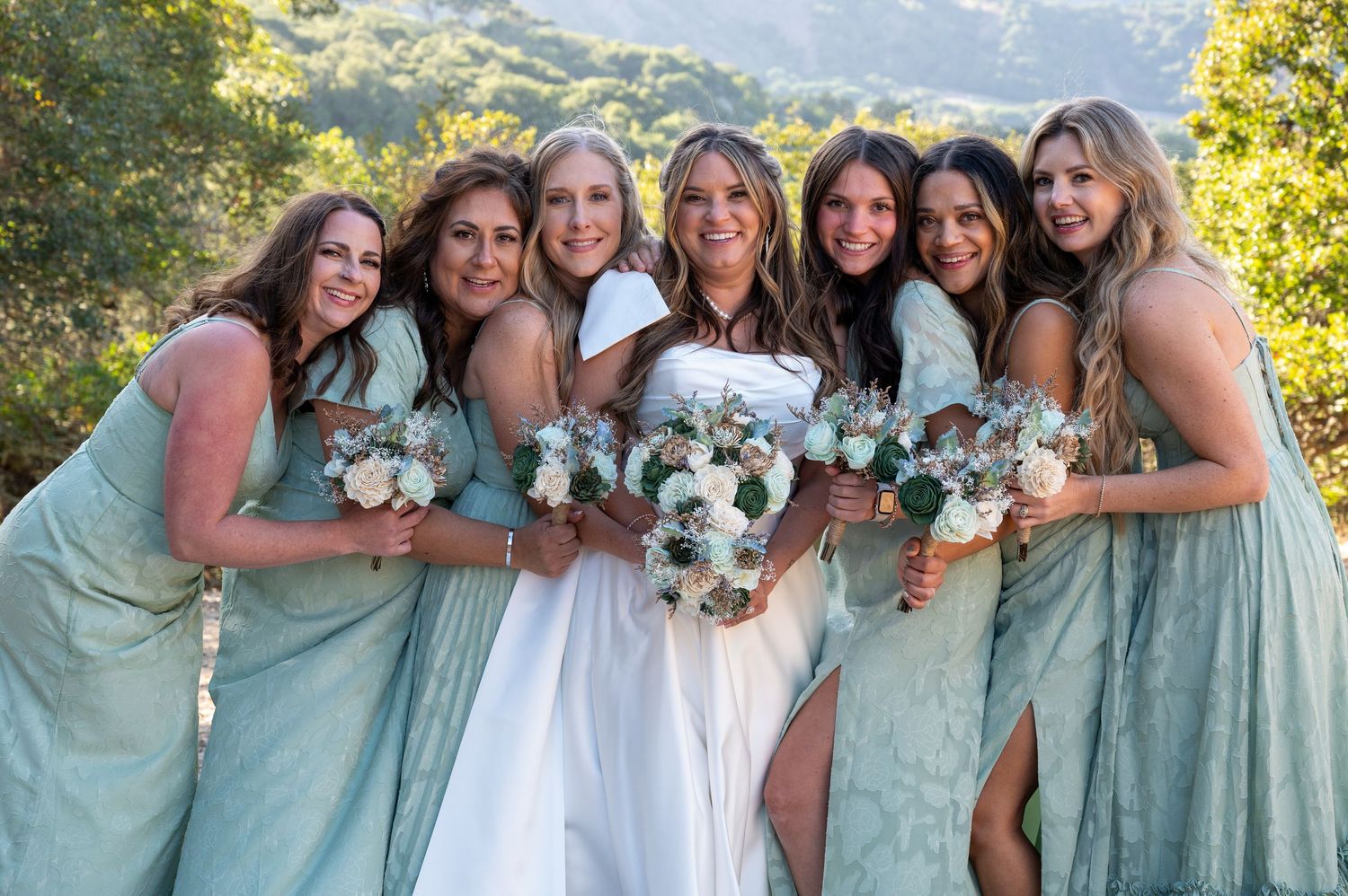 Bridesmaids in mint green dresses pose together with the bride in a white gown against a scenic outdoor backdrop.