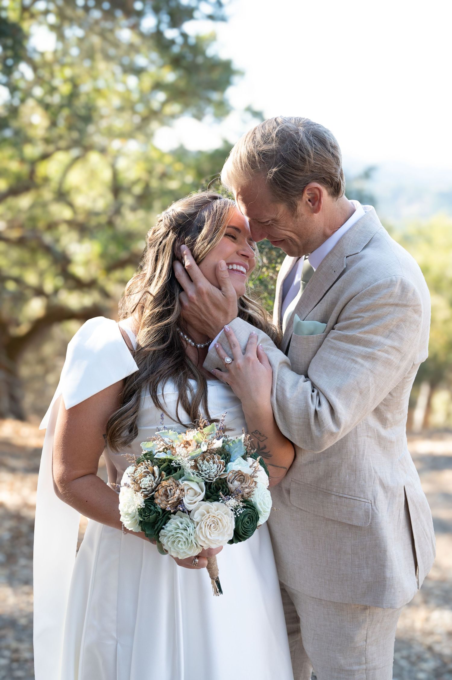Newlywed couple shares intimate moment outdoors with bride holding white and green floral bouquet.