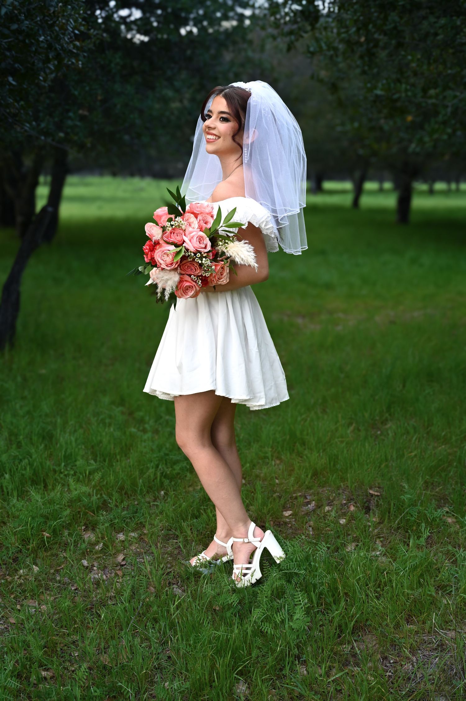Bride in short white dress and veil poses playfully in a garden setting holding coral and pink bouquet.