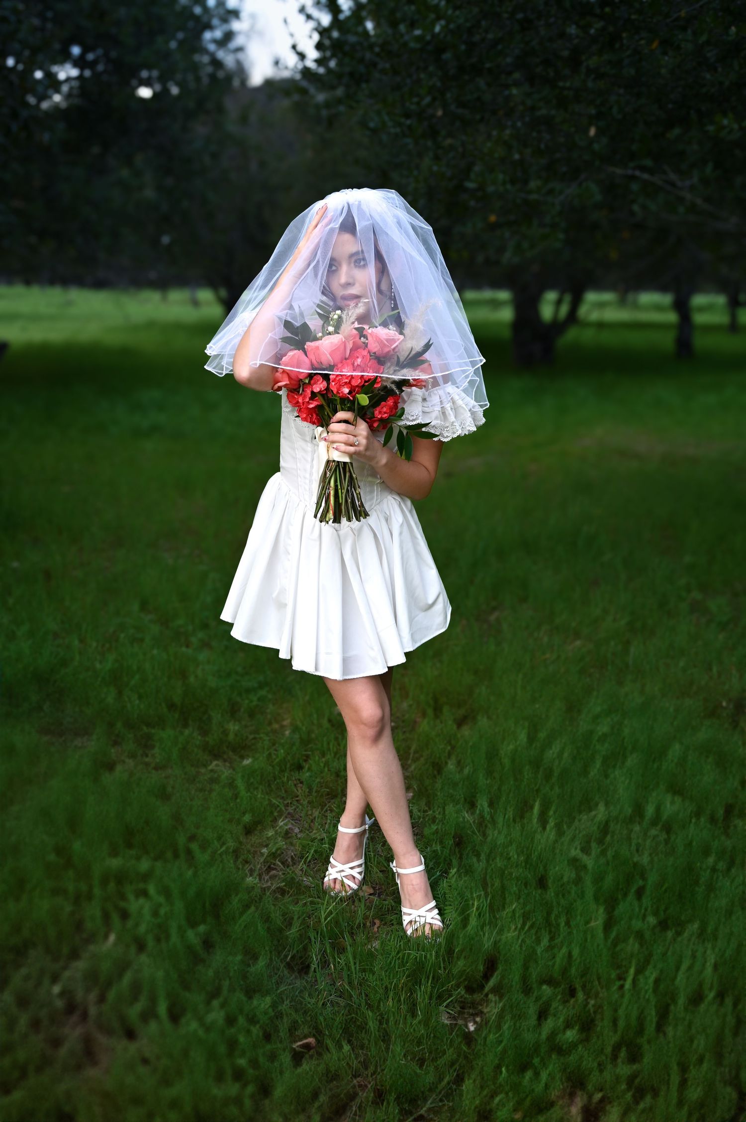Bride holding red bouquet in white dress standing in grassy field with veil flowing.