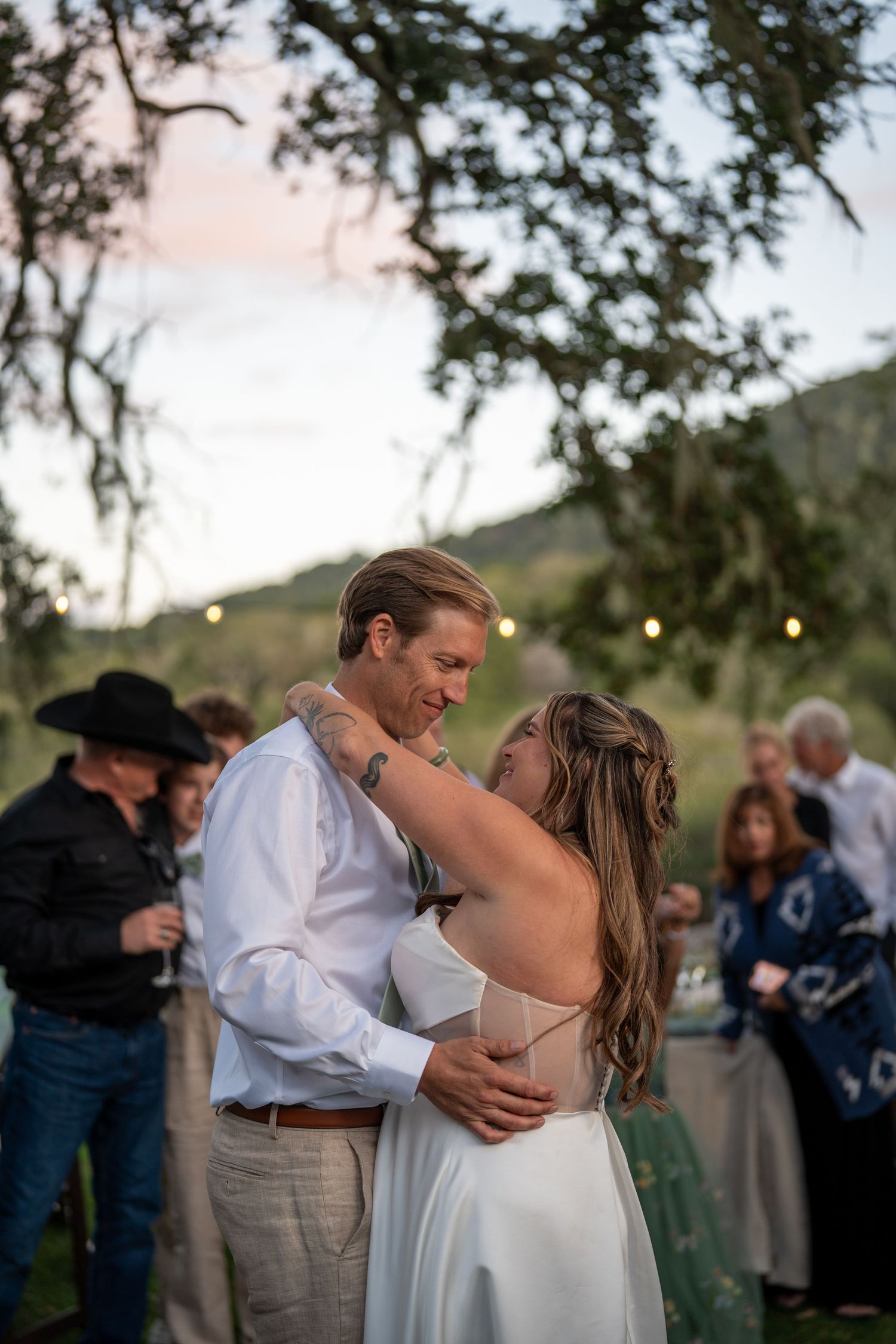 A couple shares a romantic dance outdoors at sunset with string lights and trees in the background.