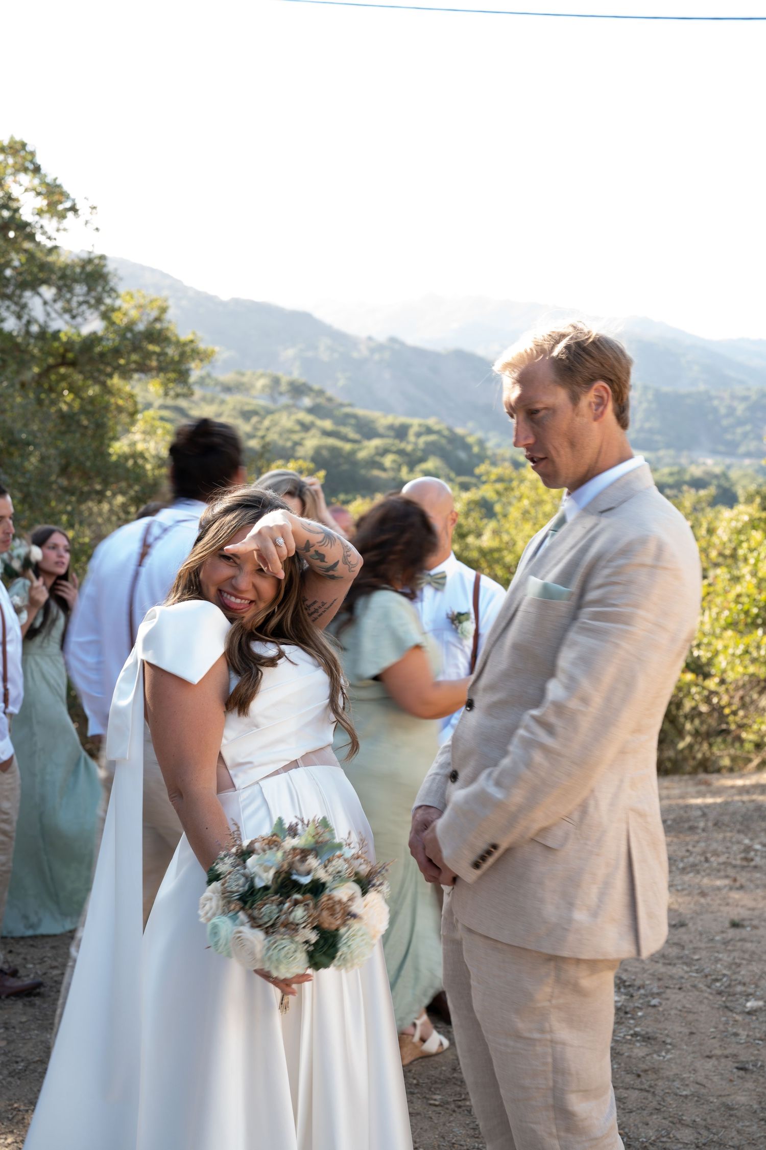 Wedding party in white dresses and tan suits embracing outdoors with mountain backdrop.
