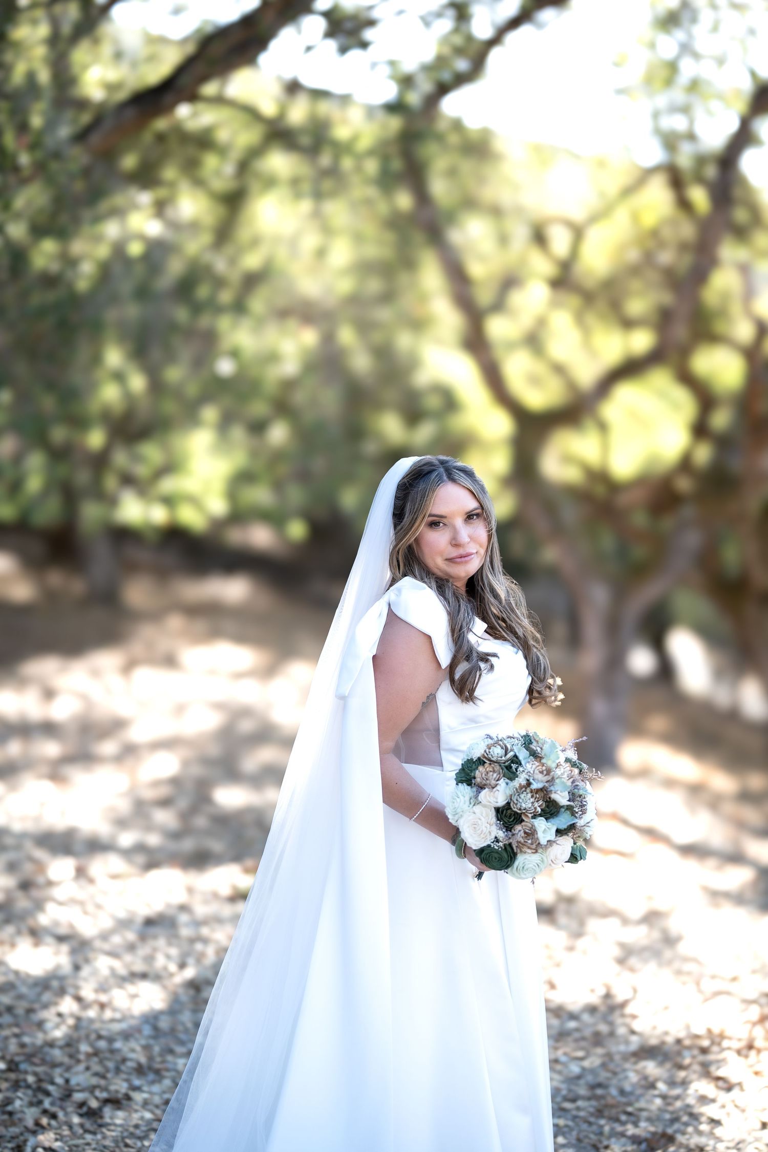 Carmel Valley Wedding A bride in a white gown and long veil holds a bouquet of white and blue flowers in a sunlit outdoor setting.
