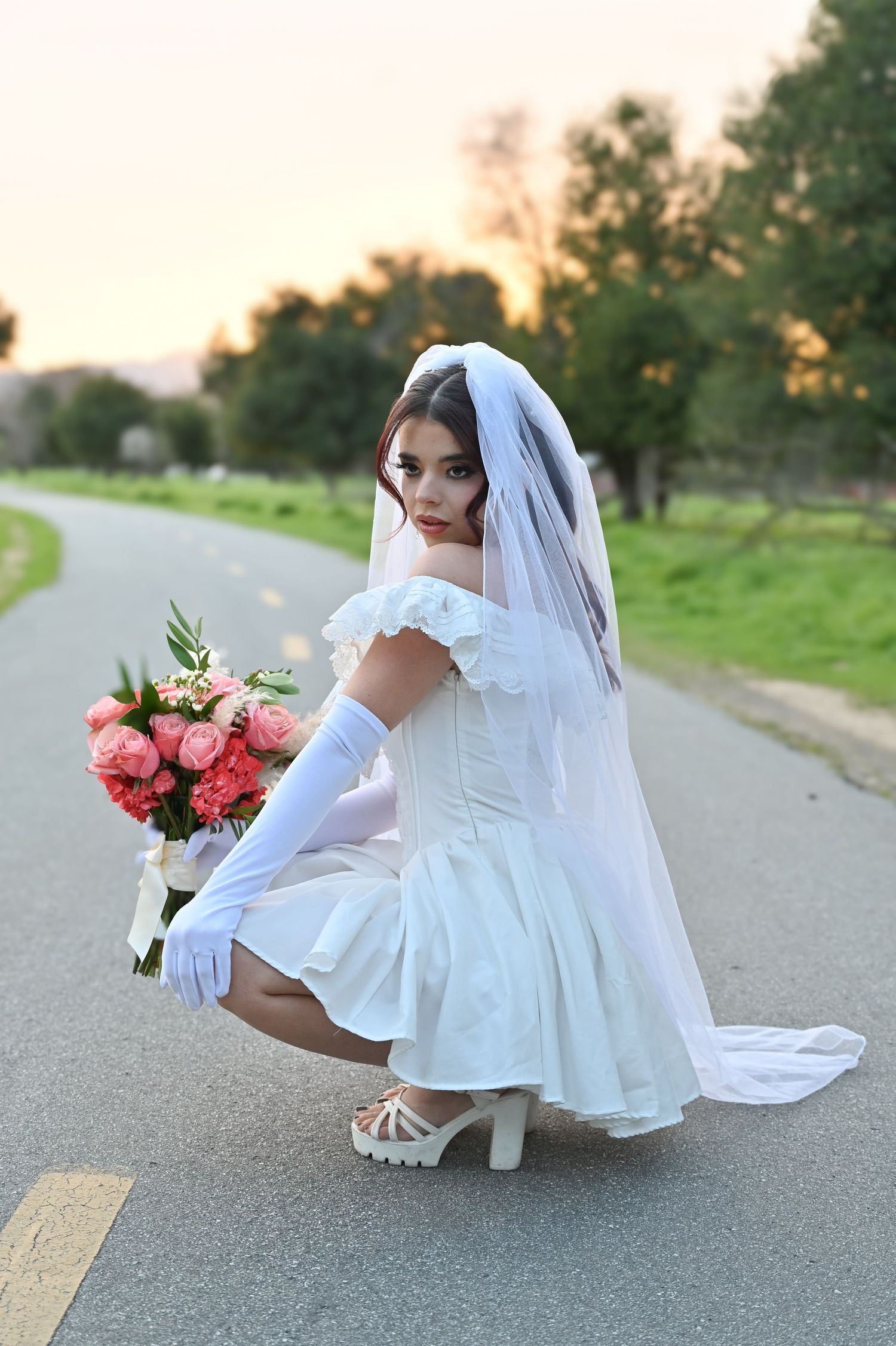 Bride in short white dress crouches on path at sunset holding pink bouquet.