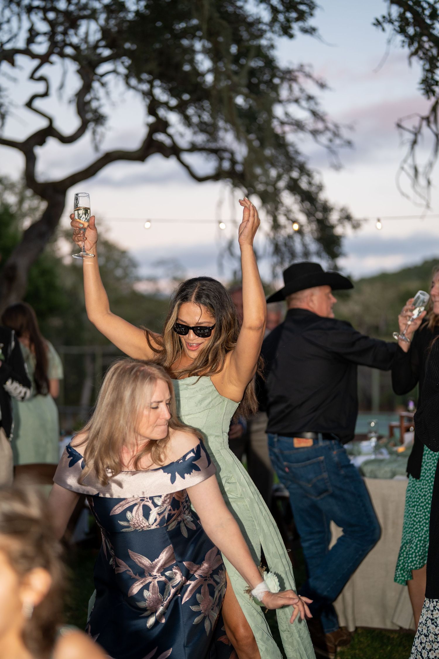 People dancing and celebrating at an outdoor party under trees at dusk with drinks in hand.