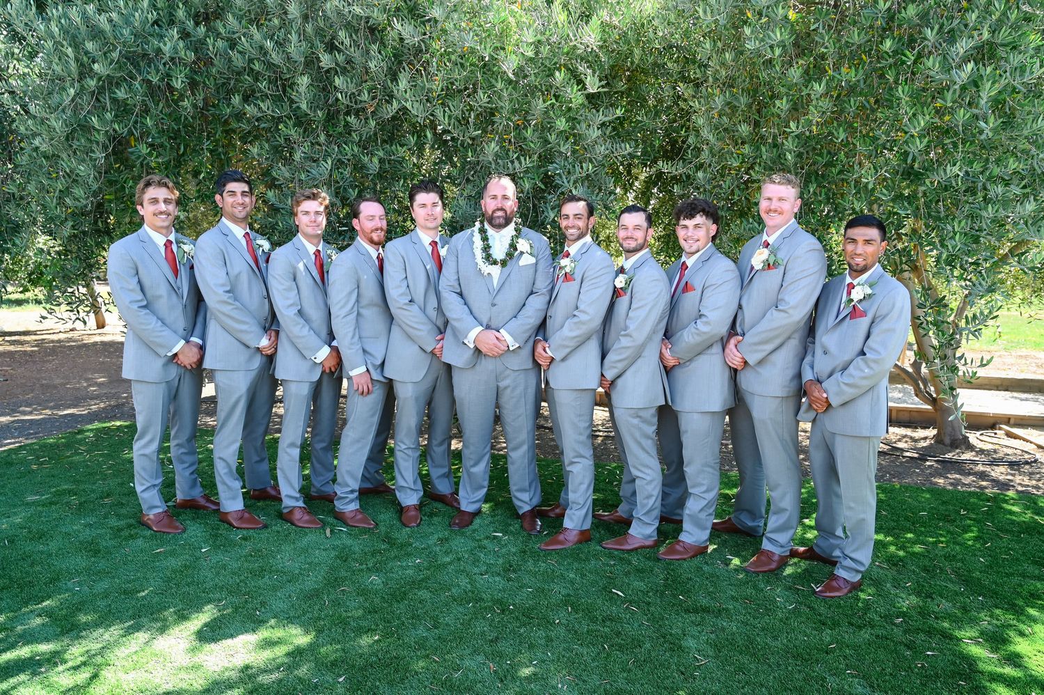 Wedding party members in gray suits pose together outdoors against lush greenery.